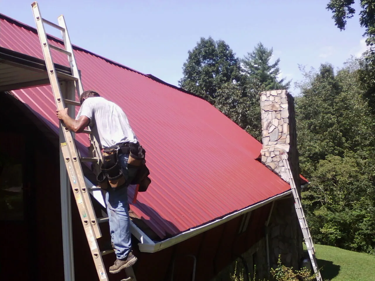 Roofer on a ladder beside a red metal roof and stone chimney — Soffit & Fascia Repair by Roofing Brothers in Glenolden